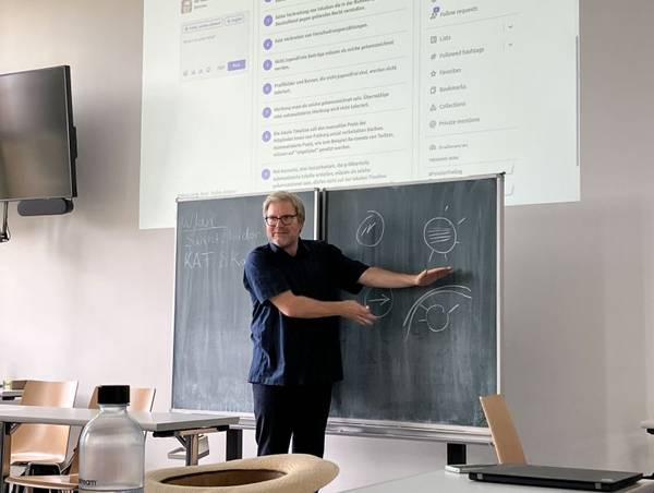Quelle: Katholische Akademie der Erzdiözese Freiburg Ein Mann in dunkelblauer Kleidung und grauen Haaren lächelt in die Kamera und erklärt den Teilnehmenden eines Seminars etwas an der Tafel, über der Tafel erscheint die digitale Präsentation.