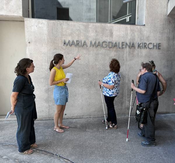 Quelle: Referat Inklusion-Generationen, Stephan Langer Blinde und sehende Menschen stehen vor dem tastbaren Schriftzug der Maria-Magdalena-Kirche in Freiburg/Rieselfeld.
